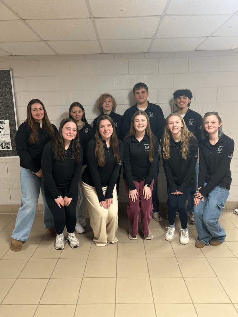 A group of teenagers crowd together to take a group photo wearing matching black jackets.