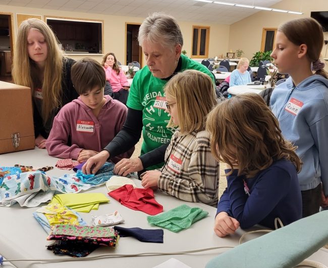 An adult 4-H volunteer works with a group of youth on picking out materials to sew with on the machine.