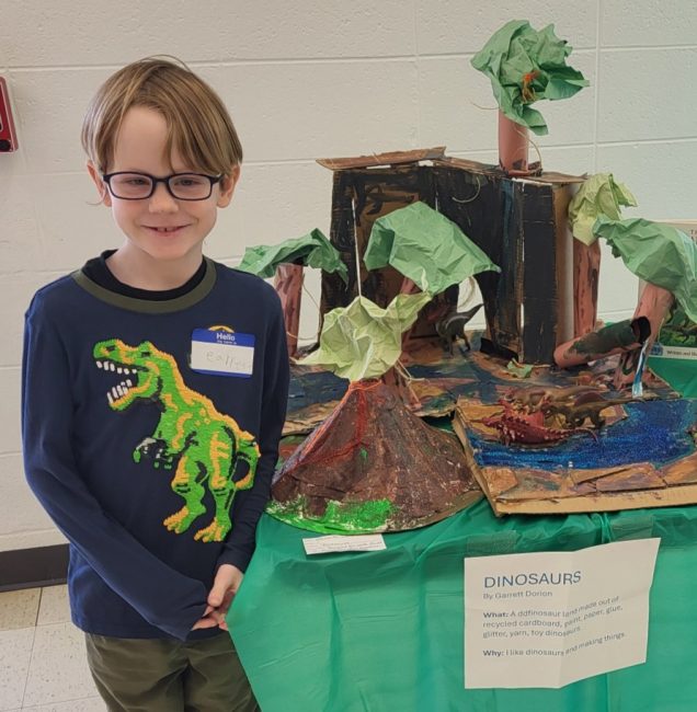 A young boy stands in front of a dinosaur art project he made for the expo.