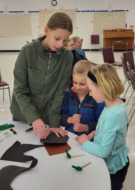 Youth in grades K-2 gather around a table and watch as a youth leader demonstrates how to do a paper craft.