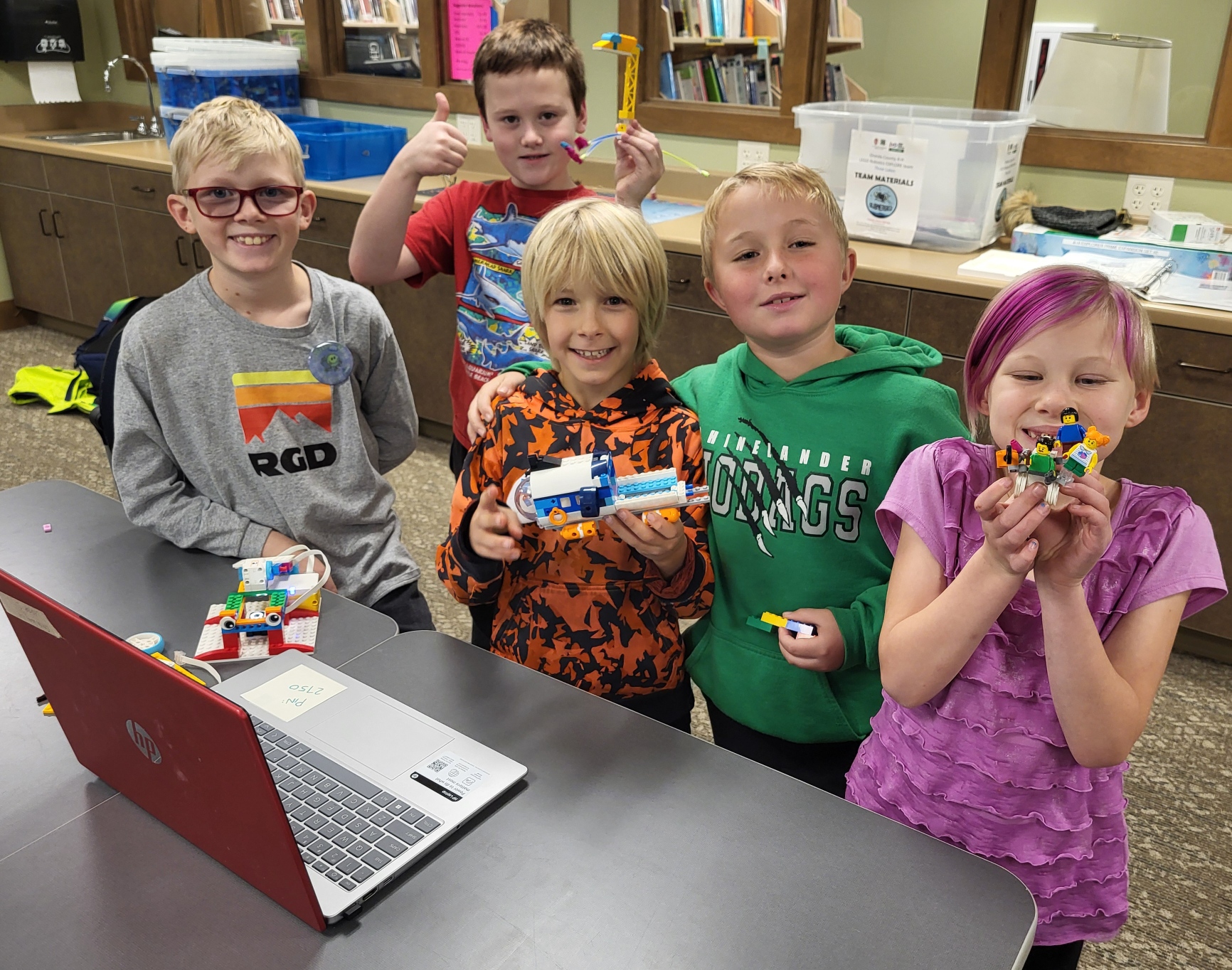 A group of young children stand in front of a laptop and hold up their Lego builds while smiling at the camera.