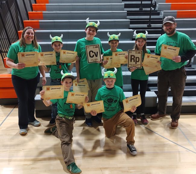 The Lego Robotics Challenge team and their two coaches stand in front of the bleachers while holding up certificates they won at the competition.