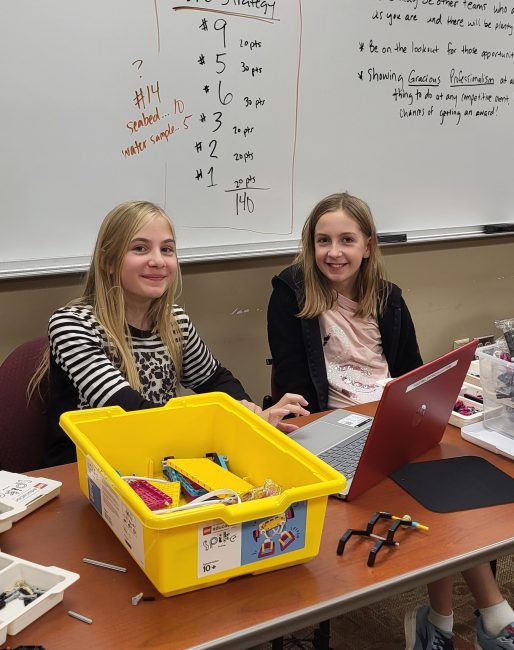 Two young girls sit side by side at a table with Legos and a laptop in front of them while they smile at the camera.
