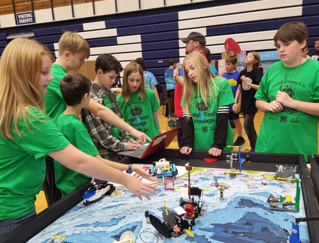 A group of youth stand around the Lego Robotics table as they work on the laptop to check their work.