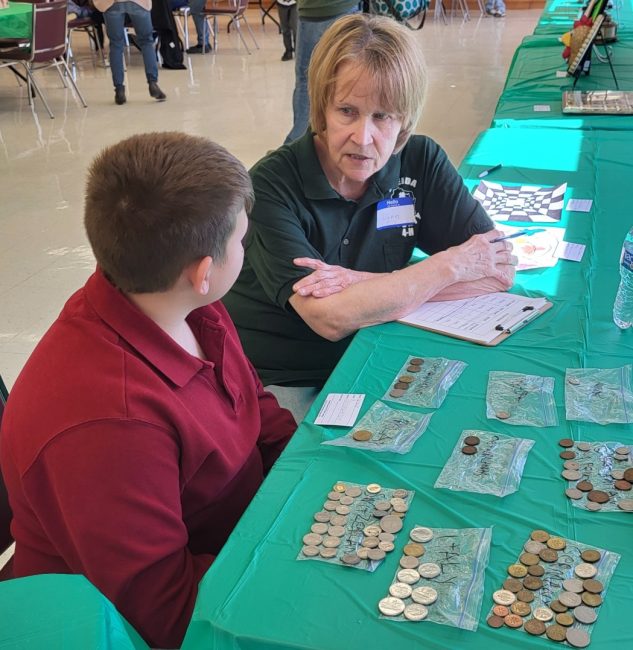 An adult volunteer sits with a youth at a long table covered with a coin collection.