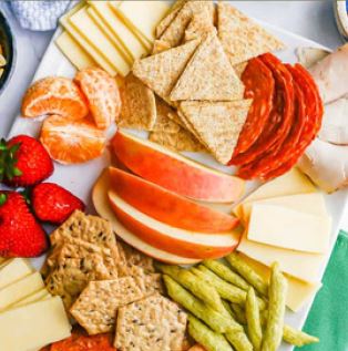 Healthy foods, such as apple slices, whole grain crackers, orange slices, and strawberries on a cutting board.