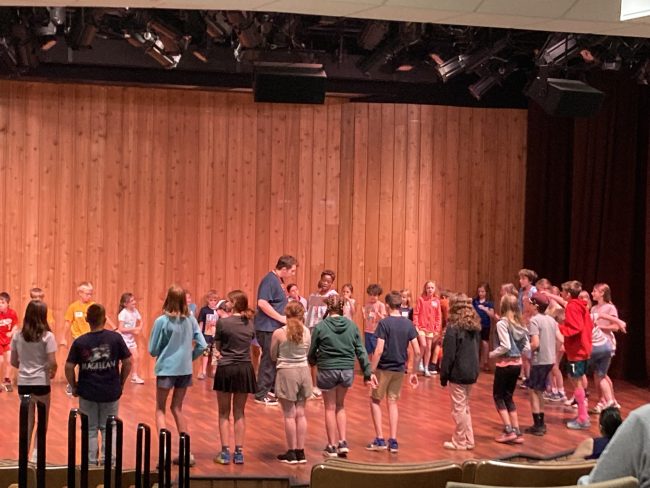 Youth circle on stage around the director and actor during a rehearsal for the musical.
