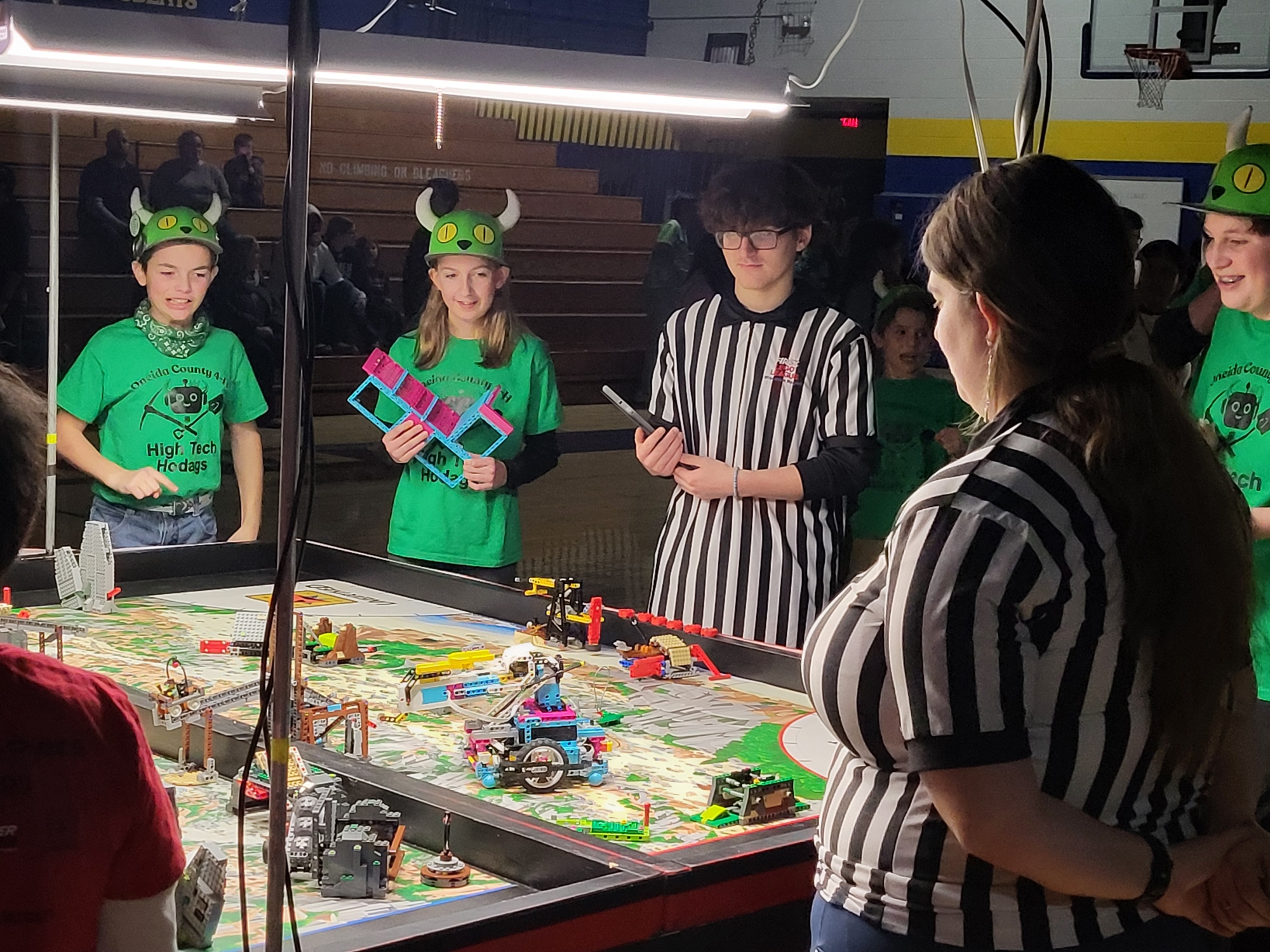 Three youth and two referees watch the Lego Robotics table as the team puts their robots into action.