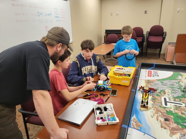 An adult coach leans over a table with three youth who are working on a Lego robotics build.