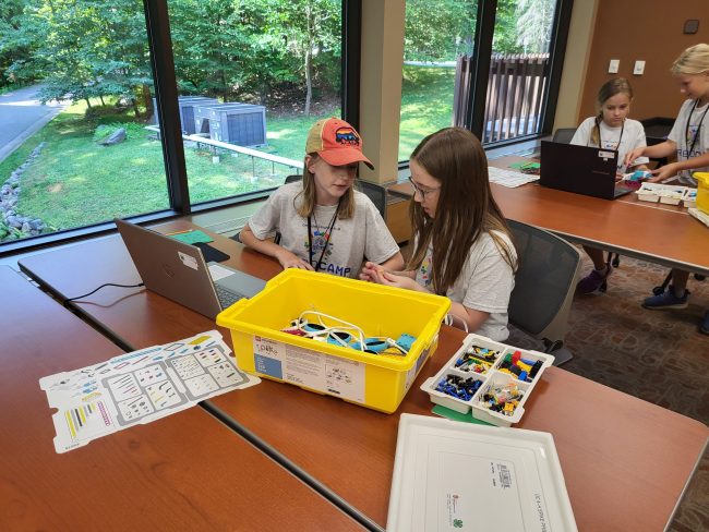 Two youth sit at a table in front of a laptop and code.