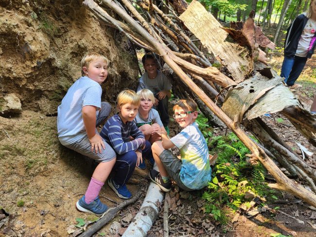 A group of youth huddle under a fort they built on Nicolet campus, composed of fallen tree logs and branches.