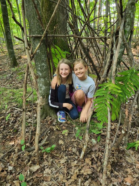 Two young girls sit outside under a large tree in the summer.