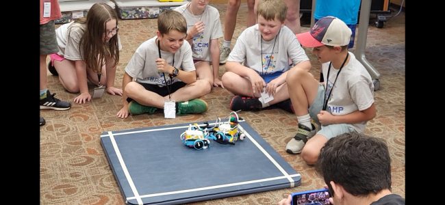 A group of youth sit on the floor in front of a small flat black tabletop and watch their robots move around.